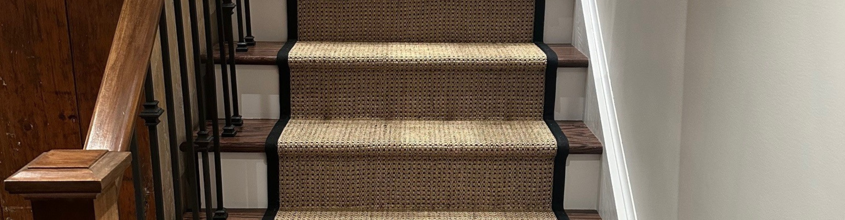A carpet runner with a black border is installed on a wooden staircase with a dark wood banister and black spindles.