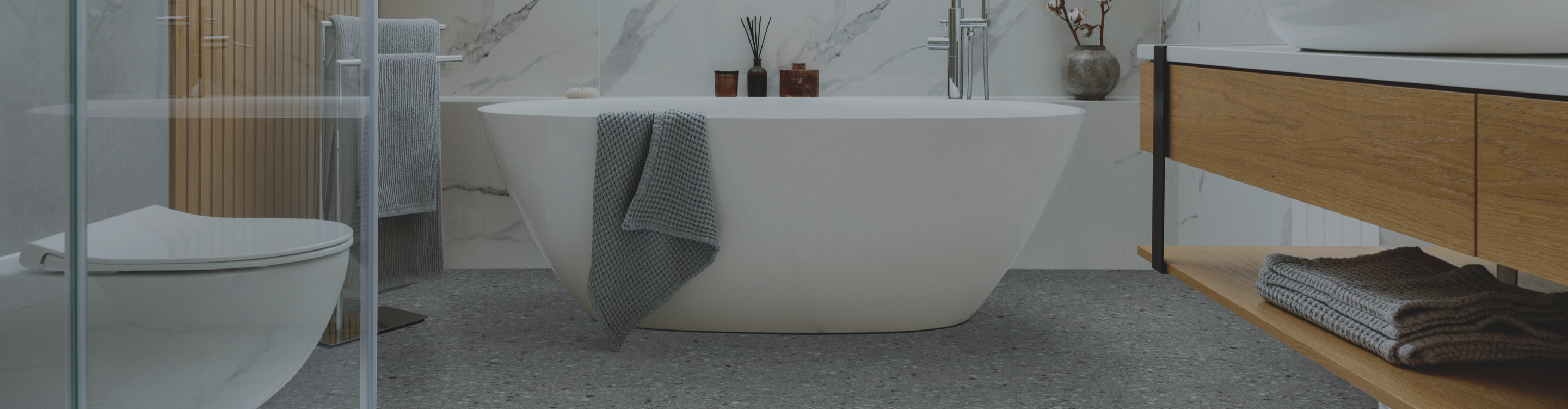 Waterproof flooring in a modern bathroom with a freestanding white bathtub, gray towels, a glass shower enclosure, a marble wall, and a wooden vanity with a countertop faucet